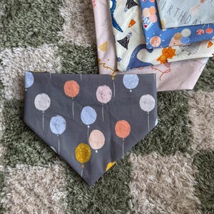 Puede incluir: Varios pañuelos para perros con temática de cumpleaños. Un pañuelo gris presenta un patrón de globos de colores. Otros pañuelos muestran texto y gráficos de cumpleaños sobre telas azul claro, rosa y estampadas. Los pañuelos están doblados.