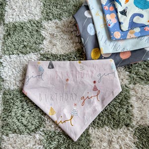 Puede incluir: Un pañuelo rosa para perro con las palabras "BIRTHDAY girl" e ilustraciones de sombreros de fiesta. Se ven varios pañuelos estampados, con globos y dinosaurios. Los pañuelos están sobre una alfombra estampada verde y blanca.