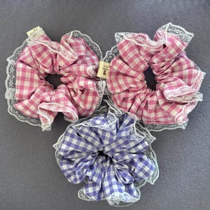 May include: Three scrunchies in pink and white, and purple and white gingham patterns. Each scrunchie is trimmed with white lace. The scrunchies are on a gray surface.