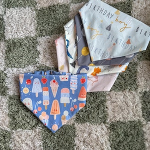 Puede incluir: Una colección de pañuelos para perros con varios diseños. Un pañuelo azul muestra ilustraciones de helados. Otros pañuelos presentan diseños de dinosaurios, globos y cumpleaños. Los pañuelos son de tela y son adecuados para mascotas.