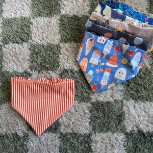 Puede incluir: Bandanas para perros con varios diseños. Una tiene rayas verticales rojas y blancas con un borde con volantes. Otras presentan diseños de helados y frutas sobre un fondo azul, y otros diseños.