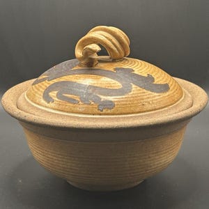 May include: A ceramic lidded bowl with a textured, light brown exterior. The lid features a dark brown dragon design and a twisted handle. The bowl is likely used for serving or storage.