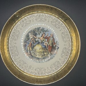 May include: A vintage decorative plate with a gold rim and a central image of a couple dancing in a garden. The plate features a cream-colored background with gold filigree patterns. The dancers are dressed in period clothing.