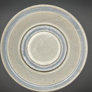 Könnte beinhalten: Eine Keramikschale mit gesprenkeltem, hellgrauem Innenraum und hellblauem Rand. Die Schale hat ein kreisförmiges Design mit erhabenem Mittelteil und einem dunkelgrauen Außenring. Die Schale steht vor einem dunklen Hintergrund.