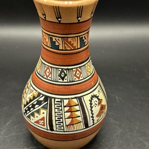 May include: A hand-painted ceramic vase with a bulbous body and a narrow neck. The vase features geometric patterns in shades of brown, orange, black, white, and yellow. The design includes stylized figures and shapes, creating a unique decorative piece.