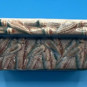 Könnte beinhalten: Rechteckiger Keramik-Pflanztopf mit einem strukturierten, blattartigen Muster. Der Pflanztopf hat eine braune Basis mit weißen und tealfarbenen Akzenten. Der obere Rand hat ein bambusartiges Design.