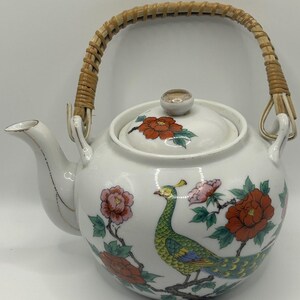 May include: White ceramic teapot with a woven handle and a lid. The teapot is decorated with a peacock and floral design in red, pink, green, and yellow. The spout and handle attachments have gold accents.