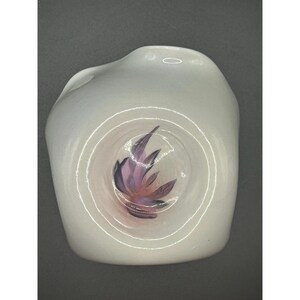 Può includere: Un vaso di vetro bianco di forma organica con un design circolare centrale. Il design presenta un motivo floreale viola e rosa. Il vaso ha una finitura liscia e lucida su uno sfondo grigio scuro.