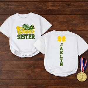 May include: Two white short-sleeve baby bodysuits. The left one features a green pom-pom, a yellow megaphone, and the text "Cheer SISTER" in yellow and green. The right one displays a yellow bow and the name "JAELYN" in green, with a gold medal.