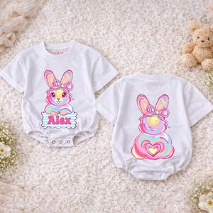 Puede incluir: Body de bebé blanco con un diseño de conejito colorido. La parte delantera presenta un conejito con un lazo y el nombre "Alex". La parte trasera del body tiene un diseño de conejito a juego. El body es de manga corta y tiene cierres a presión.