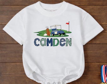 May include: White baby romper featuring a golf-themed illustration. The design includes a blue golf cart, golf bag, and the name "Camden" in blue and green patterned letters. A red flag and golf balls complete the design.