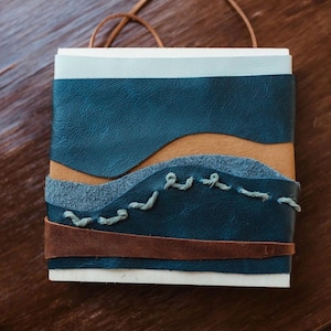 May include: A square leather-bound journal featuring layered panels of blue, tan, and brown leather. The cover includes a textured blue suede section with decorative light blue stitching and a thin brown leather strap across the bottom.