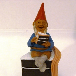 May include: A whimsical garden gnome figurine wearing a red hat and blue shirt, holding a slice of chocolate cake. The gnome is seated on a multi-layered chocolate cake with white frosting. The figurine has a smiling expression.
