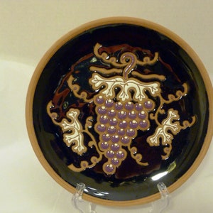 Puede incluir: Un plato de cerámica decorativo con un diseño en relieve de una vid con uvas moradas y hojas blancas sobre un fondo negro.