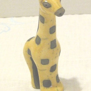 May include: A ceramic giraffe figurine with a yellow body and dark gray spots. The giraffe has a long neck, small ears, and a gray snout. The figurine is standing upright, showcasing its legs and feet.
