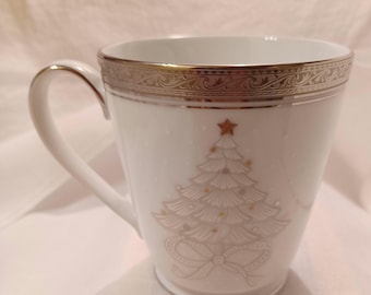 May include: White ceramic mug with a Christmas tree design. The mug features a gold rim and handle, and a detailed illustration of a Christmas tree with a star on top. The design includes a bow at the base of the tree.