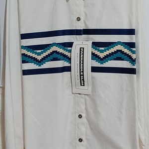 Peut inclure: Chemise boutonnée à manches longues blanche avec un motif géométrique bleu et turquoise sur la poitrine. La chemise porte une étiquette indiquant "PANHANDLE SLIM QUALITY WESTERN WEAR".