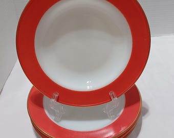 May include: A stack of white and red rimmed plates with a gold trim. The plates are stacked on a clear plastic plate stand. The plates have a white center and a red border.