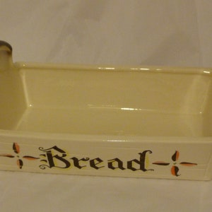 May include: A white ceramic bread basket with brown handles and the word "Bread" written in black and yellow lettering. The basket has a decorative design on the sides.
