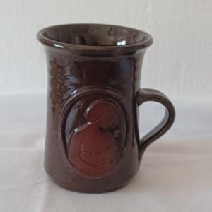 Może przedstawiać: Brązowy kubek ceramiczny z wypukłym wzorem przedstawiającym kobietę trzymającą dziecko. Kubek ma uchwyt i jest pokryty błyszczącą glazurą.