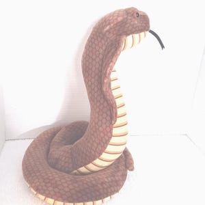 May include: A plush brown cobra snake toy with a detailed scale pattern. The snake is posed upright, with its head raised and tongue extended. The underside of the neck features cream and tan stripes.