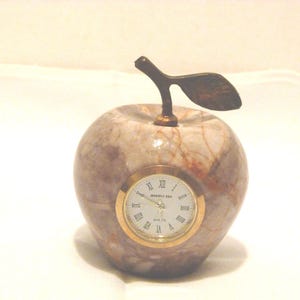 May include: A polished marble apple-shaped desk clock featuring a brown and white marbled pattern. The clock face is circular with a gold-toned rim, white background, black Roman numerals, and black hands. A brass-colored metal stem and leaf sit on top.