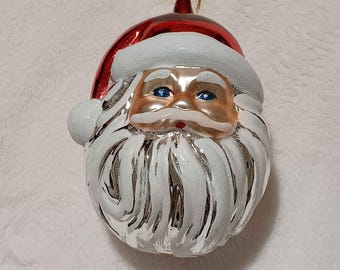 May include: A Christmas ornament shaped like Santa Claus's head. The ornament has a red hat, a white beard, and a gold face. The eyes are blue, and the beard has a silver glitter effect. A gold-colored loop is at the top for hanging.