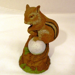 May include: A detailed figurine of a chipmunk holding a golf ball. The chipmunk is brown with black stripes and is perched on a tree stump base. The golf ball is white, and the base has green leaves.