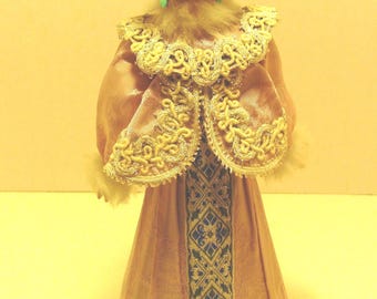 May include: A decorative doll wearing a light pink gown with a central blue and white geometric patterned panel, a matching bolero jacket with gold trim, and a large, fluffy white faux fur hat with dangling green teardrop earrings.