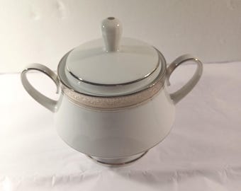 May include: White porcelain sugar bowl with lid and two handles. The bowl has a silver trim around the rim and base, and a decorative band. The lid has a small knob on top. The sugar bowl is designed for serving sugar.