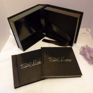 Puede incluir: Caja de regalo negra con un juego de libros pequeños a juego. La caja está abierta, revelando un interior brillante y una cinta negra. Los libros tienen un patrón de rayas y la firma "Frank Sinatra" en plata.