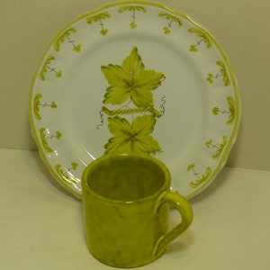 以下が含まれることがあります： 緑と茶色の葉のデザインと波打つ縁のある白いセラミックプレート。取っ手付きの明るい黄色のセラミックマグがプレートの上に置かれています。