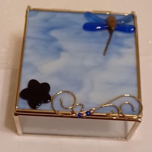 Puede incluir: Una pequeña caja de vidrio con un diseño de mármol azul y blanco. La caja tiene un borde dorado y presenta una libélula, una estrella y un diseño de espiral.