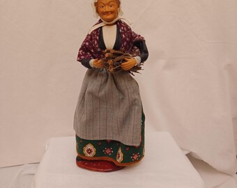May include: A handcrafted figurine of an elderly woman, dressed in traditional attire. She wears a white bonnet, a patterned shawl, and a striped skirt. The figurine is holding a bundle of sticks, with a red and green patterned base.