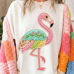 May include: White t-shirt featuring a colorful flamingo applique. The flamingo has a pink body, a turquoise wing with floral details, and a yellow beak. The design is outlined with white beads. The sleeves of a multi-colored sweater are visible.