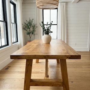 Könnte beinhalten: Ein großer, rechteckiger Esstisch aus Holz mit natürlicher Oberfläche. Der Tisch hat ein stabiles Gestell im Stil einer Bockkonstruktion und steht in einem hellen Raum mit großen Fenstern und einer gewebten Pendelleuchte. Eine Vase mit Grün steht auf dem Tisch.