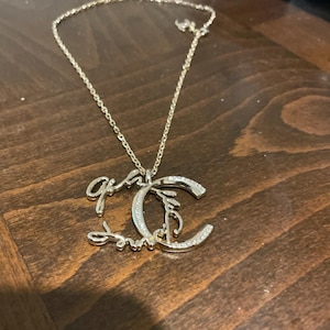 以下が含まれることがあります： 「Gia」と「Smith」の筆記体と、様式化された「C」の形をしたペンダントが付いたシルバーカラーのネックレス。ペンダントには、小さな輝く石が飾られています。ネックレスチェーンは繊細で、シルバーカラーです。