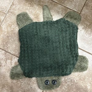 May include: A green turtle-shaped bath mat with a textured, rectangular body and four flipper-like appendages. The turtle's head features two black button eyes. The mat is designed to resemble a turtle.