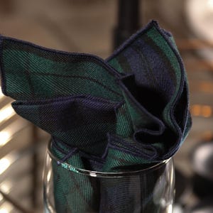 Peut inclure: Un carré de poche à carreaux vert et bleu marine plié, présenté dans un verre transparent. Le tissu présente un motif tartan classique, avec des lignes entrecroisées vert foncé et bleu marine. Le carré de poche est soigneusement disposé.