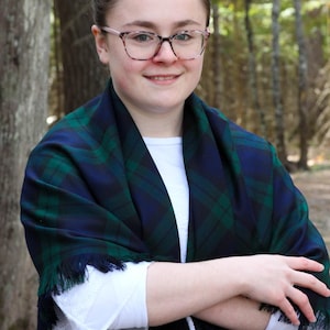 May include: A green and navy blue plaid shawl with fringe. The shawl is draped over a white long-sleeved shirt. The person is wearing glasses with a light pink frame.