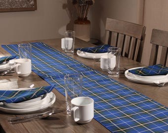May include: A blue plaid table runner with thin yellow and white stripes laid across a wooden table. The table is set with white ceramic plates, white mugs, clear drinking glasses, and folded blue plaid napkins matching the table runner pattern.