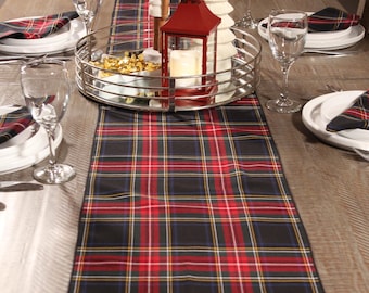 May include: A festive table setting with a red and black plaid table runner. The centerpiece includes a red lantern, a white ceramic Christmas tree, and a silver serving tray. Wine glasses and plates with plaid napkins complete the holiday decor.