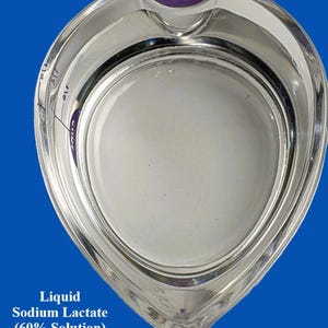 Op de afbeelding: Een heldere glazen maatbeker gevuld met een melkachtige witte vloeistof, met het label "Liquid Sodium Lactate (60% Solution)". De beker heeft een gebogen handvat en maatstreepjes. De achtergrond is effen blauw.