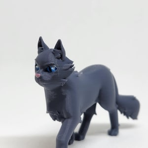 Puede incluir: Figura impresa en 3D de un gato en posición de caminar. El gato es de color gris oscuro con ojos azules y nariz rosa. La figura tiene una superficie texturizada y está sobre un fondo blanco. Objeto decorativo.