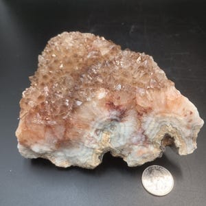 May include: A natural, rough-cut crystal formation with a mix of brown, tan, and white hues. The crystal has a textured surface with visible crystalline structures. A US quarter coin is placed next to the crystal for size comparison.
