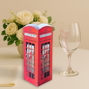 Könnte beinhalten: Ein rotes, dreidimensionales Papiermodell einer britischen Telefonzelle mit dem Wort TELEPHONE auf den oberen Paneelen. Die Struktur verfügt über ein gitterartiges Fensterdesign auf jeder Seite, das eine gedruckte Innenansicht der Telefonzelle zeigt.