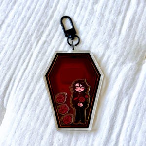 May include: A coffin-shaped acrylic keyring featuring a dark red interior with a black border. Inside, a cartoon character with dark hair and red eyes stands next to three red roses. The charm is attached to a black metal lobster claw clasp.