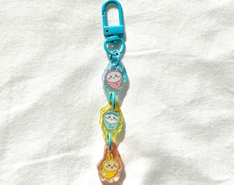May include: A blue keychain with three colorful, cartoon character charms. The charms are in the shape of irregular ovals and feature cute animal designs in pink, yellow, and blue. The keychain has a blue clasp and connecting rings.