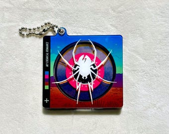 May include: A square acrylic keychain featuring a white spider graphic centered over a circular design with pink, blue, and red desert landscape colors. The text MY CHEMICAL ROMANCE is printed vertically on the left side with a small plus sign below.