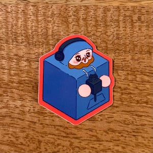 May include: A sticker featuring a cartoon character inside a blue box with headphones. The character has a brown beard and is wearing a blue hat. The box has a black cat on the front and is outlined in red.